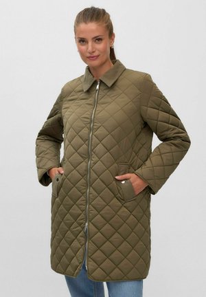 Femme portant une longue veste matelassée vert olive avec une fermeture éclair sur le devant et les mains dans les poches à boutons-pression.