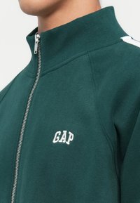 Green zip-up sweatshirt with a high collar, featuring a white embroidered "GAP" logo on the left chest and textured fabric.