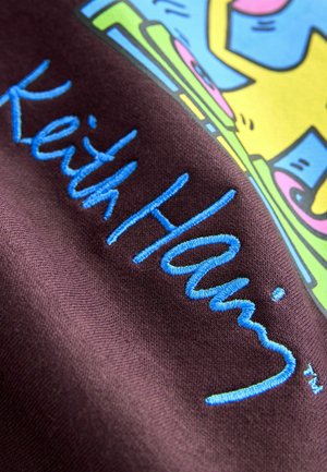 Lila tyg med broderad blå text som säger "Keith Haring." Bakgrunden har färgglada grafiska designelement i gult, rosa och grönt.