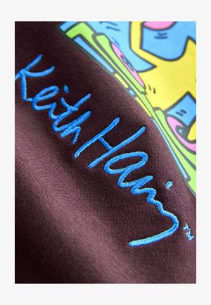 Lila Stoff mit gesticktem blauen Text "Keith Haring". Der Hintergrund zeigt bunte grafische Designelemente in Gelb, Pink und Grün.