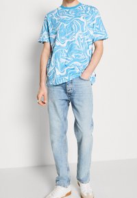 Uomo che indossa una maglietta a spirale blu chiaro e bianca, jeans blu chiaro, scarpe da ginnastica bianche, con la mano destra in tasca e la sinistra rilassata.