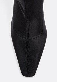Czarny botek na wysokim obcasie z wydłużonym noskiem i gładkim satynowym wykończeniem. Posiada widoczne szwy i dopasowany krój przy kostce.