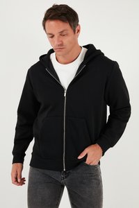 Crna zip-up hoodie s kapuljačom, napravljena od mekog materijala. Ima prednje džepove i rebraste rukave, nosi se preko bijele majice.
