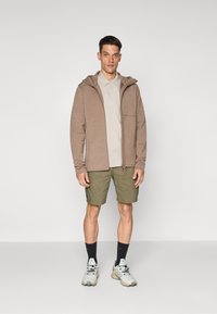 Felpa marrone con zip e cappuccio, camicia button-up grigia chiara, pantaloni cargo verdi, calze scure e sneakers chiare. Outfit casual da esterno.