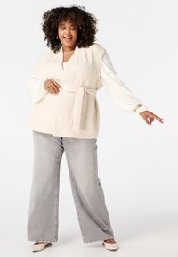 Beige gewatteerde vest met een strikceintuur, gelaagd over een witte, textuurrijke top met lange mouwen, gecombineerd met grijze, wijde jeans en lichte schoenen.