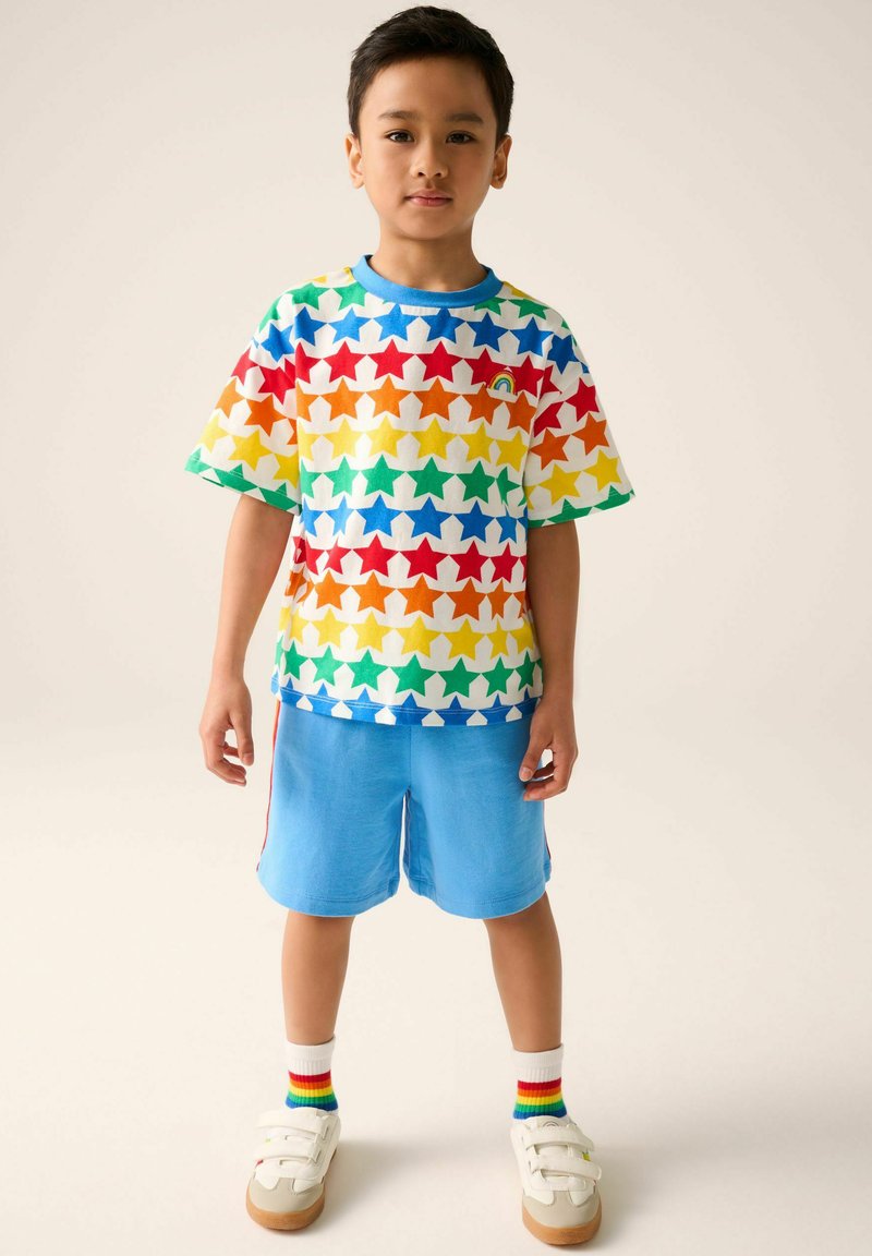Camiseta de manga corta colorida con un patrón de estrellas en rojo, azul, verde, amarillo y naranja; combinada con pantalones cortos azul claro y zapatillas blancas.
