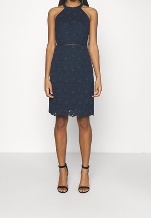 Frau trägt ein ärmelloses, marineblaues Spitzenkleid mit Neckholder und schwarze High-Heel-Sandalen mit Knöchelriemen.