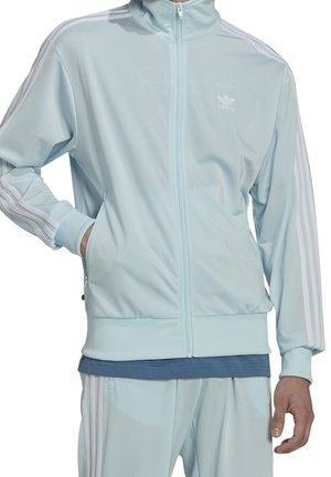 Hombre con chaqueta deportiva Adidas azul claro con rayas blancas en las mangas y pantalones a juego, con la mano en el bolsillo, de pie contra un fondo blanco.
