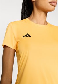 Żółta sportowa koszulka z krótkim rękawem wykonana z gładkiej tkaniny, z czarnym logo z trzema paskami na lewym ramieniu i płaskimi szwami.