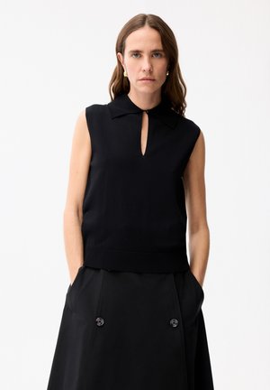 Žena s dlouhými hnědými vlasy má na sobě černý bezrukávový top s límečkem a černou sukni s knoflíky, ruce v kapsách, na bílém pozadí.