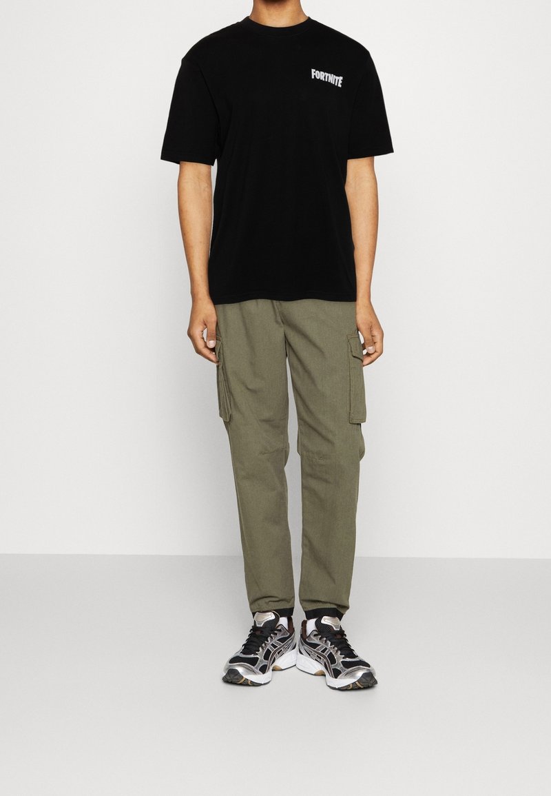 Person iført en sort Fortnite t-shirt, olivengrøn cargo-bukser og sort-hvide sportssko foran en ensfarvet baggrund.