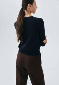 Maglione blu navy con maniche lunghe e scollo rotondo, abbinato a pantaloni marroni su misura. Tessuto morbido e setoso con dettagli minimi.
