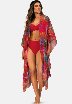 Kobieta ubrana w czerwone bikini i kolorową, zwiewną narzutkę plażową z abstrakcyjnymi wzorami, stojąca boso na jednolitym tle.