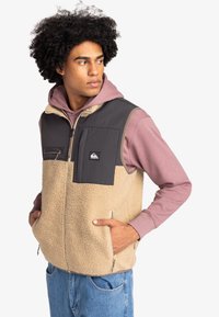 Beige Fleeceweste mit grauem Nylon-Oberteil, Reißverschluss auf der Vorderseite, zwei Seitentaschen und einer Brusttasche, getragen über einem rosa Hoodie.