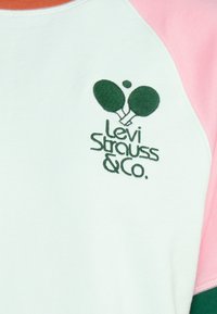 Lyhythihaisessa, pyöreäkaulaisessa collegepaidassa on mintunvihreä kangas, vaaleanpunaisia ja oransseja yksityiskohtia sekä vihreä brodeerattu Levi Strauss & Co. -logo.