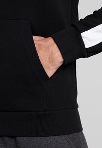 Sudadera negra con un bolsillo canguro, puños de punto y una franja blanca a lo largo de la manga para contraste. Textura de tela suave.