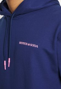 Sudadera azul marino hecha de una tela suave con un bolsillo tipo canguro. Presenta el logo rosa "SCOTCH & SODA" y extremos de cordón rosa a juego.