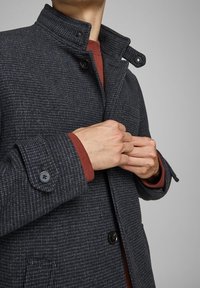 Mörkgrå texturerad ullkappa med ståkrage, med knappstängning och kontrasterande röda inre ärmslut.