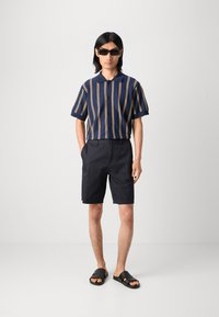 Marineblå og gulstripet kortermet skjorte med fem knapper; kombinert med svarte shorts og svarte glidesandaler med teksturerte stropper.
