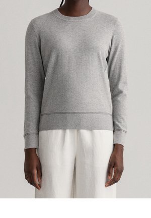 Personne portant un pull à manches longues gris clair uni et un pantalon blanc ample, visible du cou jusqu’au milieu de la cuisse sur un fond uni.