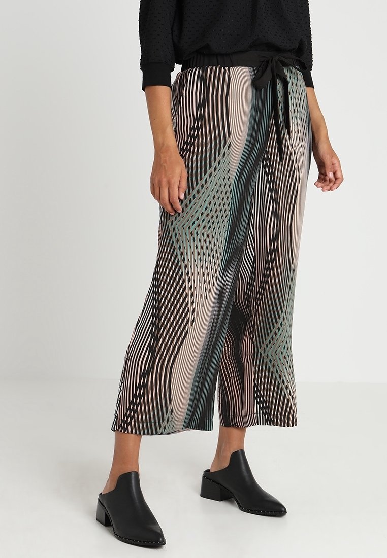 Femme portant un pantalon taille haute à jambes larges avec des rayures abstraites noires, beiges et vertes et des chaussures à talons en cuir noir faciles à enfiler.