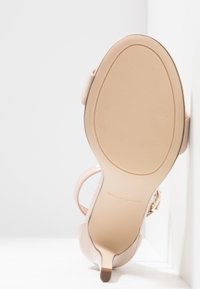 Sandale à talon haut nude avec une finition en vernis lisse, une sangle fine autour de la cheville et un talon effilé ; semelle en caoutchouc texturé pour une bonne adhérence.