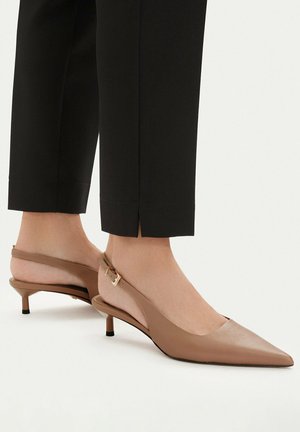 Stopy w beżowych czółenkach na niskim obcasie typu slingback z ostrym noskiem, zestawione z czarnymi spodniami o skróconej długości z rozcięciami przy kostkach.