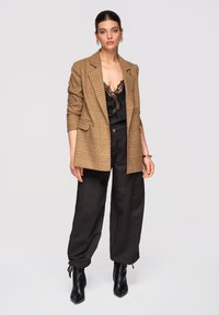 Kvinde iført brun ternet blazer, sort camisole med blonder, løse sorte bukser og sorte ankelstøvler, stående foran en ensfarvet baggrund.