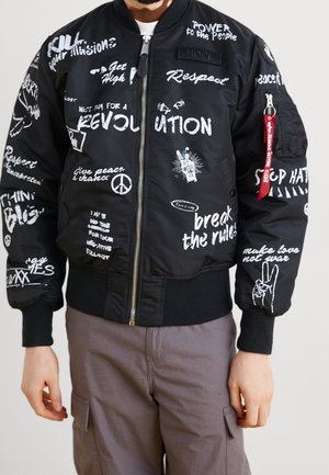 Veste bomber noire avec des slogans d'activistes blancs comme « Révolution », « Respect » et « Faites l’amour pas la guerre », portée par une personne du cou aux hanches.