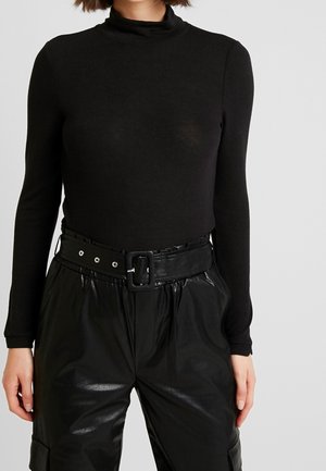 Femme portant un haut noir moulant à manches longues et col roulé, et un pantalon noir en similicuir taille haute avec une large ceinture.