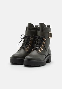 Botines de piel negra con suela gruesa, detalles en metal dorado y cordones negros. Presentan un cuello acolchado y una correa ajustable.