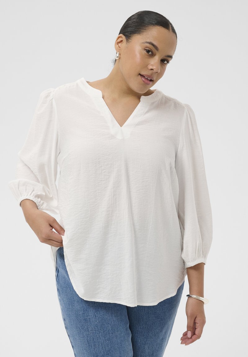 Femme aux cheveux foncés portant un chemisier blanc à col en V à manches longues et un jean bleu, posant devant un fond clair uni.