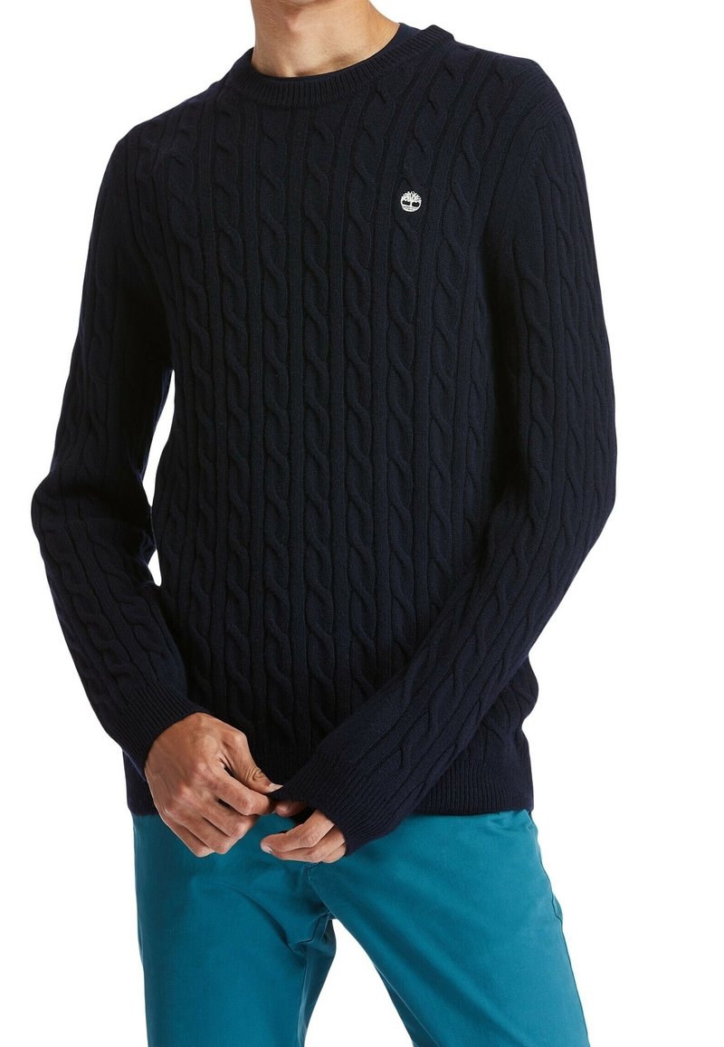 Pull tricoté bleu marine avec un motif en torsades, un col et des poignets côtelés, et un petit logo sur la poitrine. Porté avec un pantalon bleu sarcelle.