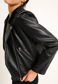 Giacca da motociclista in pelle nera con zip asimmetrica, spalline con bottoni a pressione e due tasche con cerniera. Texture liscia e design aderente.