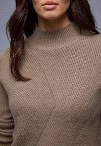 Pull en maille côtelée beige avec un col montant, présentant des motifs texturés diagonaux et des manches ajustées, mettant en valeur un matériau doux et texturé.