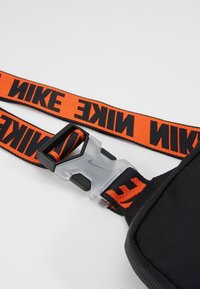 Taška Nike s černým textilním tělem, širokým oranžovým popruhem s černým textem "NIKE" a průhlednou plastovou sponou pro připevnění.