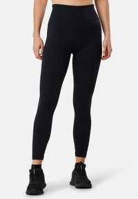 Leggings noirs taille haute en matériau extensible, pr�ésentant une texture lisse et un design ajusté, associés à des chaussures de sport noires.