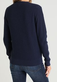 Suéter azul marino con un patrón de punto texturizado, cuello redondo y puños y dobladillo acanalados. Usado con jeans oscuros, mostrando la vista trasera.