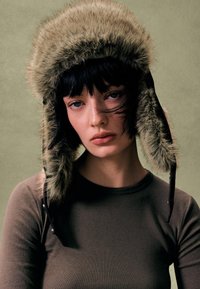 Pălărie din blană artificială verde măsliniu cu clapete pentru urechi, luciu subtil și accente din piele neagră. Purtați cu un top maro cu mâneci lungi și croială strânsă.