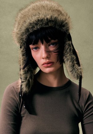 Kapelusz z futra ekologicznego w oliwkowej zieleni z nausznikami, subtelnym połyskiem i czarnymi skórzanymi dodatkami. Noszony z dopasowanym brązowym topem z długim rękawem.