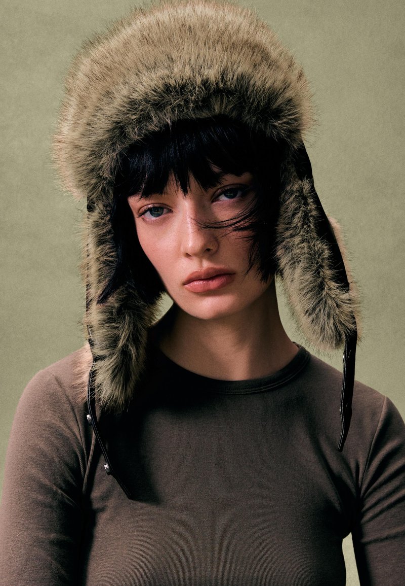 Pălărie din blană artificială verde măsliniu cu clapete pentru urechi, luciu subtil și accente din piele neagră. Purtați cu un top maro cu mâneci lungi și croială strânsă.