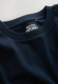 Marinblå bomullströja med ribbad rundad hals och vit "ORIGINAL SUPERDRY" text på innanför etiketten. Mjuk textur, avslappnad design.