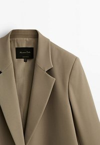 Beige Blazer aus einem strukturierten Stoff, mit Reverskragen und einem schlichten Etikett. Das Design zeichnet sich durch einen modernen Schnitt und klare Linien aus.