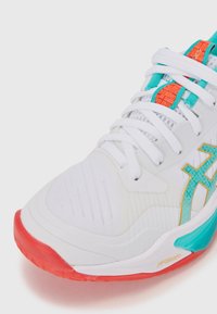 Chaussure de sport blanche avec une tige texturée, des accents turquoise et orange, et une semelle en caoutchouc rouge au design distinctif.