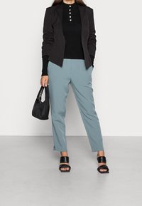 Femme portant un blazer noir, un haut noir à boutons, un pantalon bleu clair, des sandales noires, tenant un sac à main noir, debout sur un sol gris.