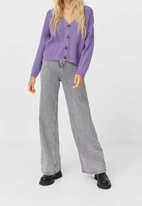 Paarse gebreide cardigan met een V-hals en knoopsluiting, gecombineerd met grijze wijde jeans en zwarte dikke laarzen.
