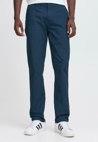 Moški nosi mornarsko modre ravne hlače in bele superge z črnimi črtami, stoji proti enostavni svetli ozadju.