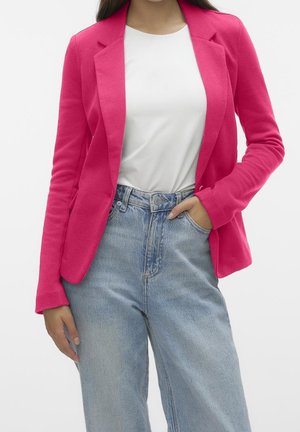 Kobieta w jasno-różowej marynarce, białym t-shircie i jasnoniebieskich wysokich jeansach, stoi z jedną ręką w kieszeni.