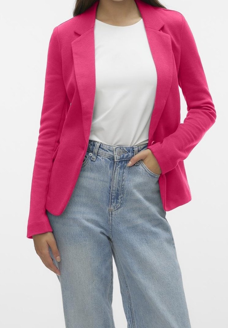 Femme portant un blazer rose vif, un t-shirt blanc et un jean bleu clair taille haute, debout avec une main dans la poche.