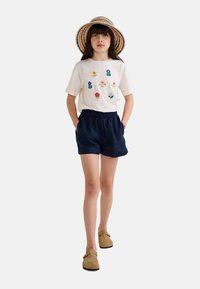 T-shirt blanche avec broderie florale colorée, associée à un short bleu marine et des chaussures slip-on marron. Un chapeau en paille multicolore complète la tenue.